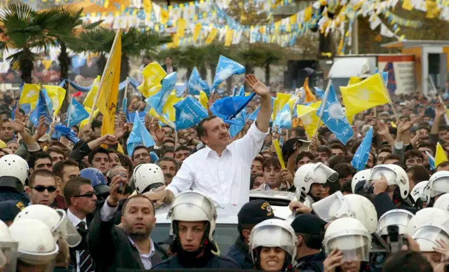 Erdoğan, 3 Kasım 2002 seçimleri öncesinde kampanya sırasında. Arabadan el sallıyor. Önünde bir grup polis var. 

