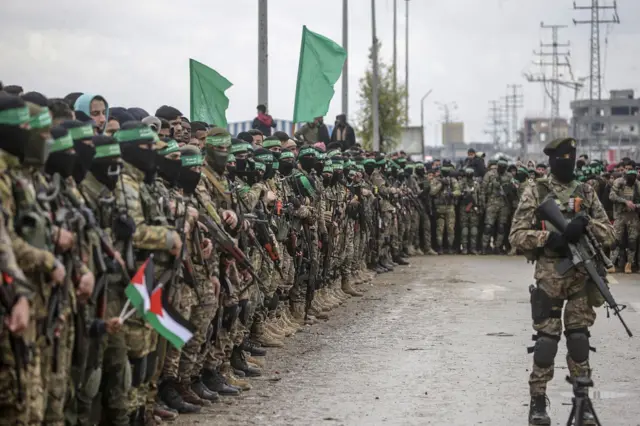 Homens fardados e fortemente armados enfileirados; é possível ver bandeiras palestinas