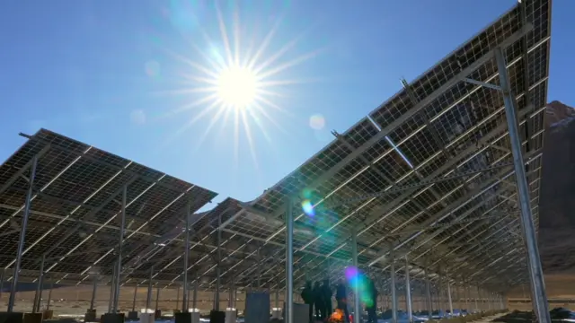 Solar panels stand tilted on tall poles under a bright, cloudless sky, with a group of people gathered in the shade beneath the panels.