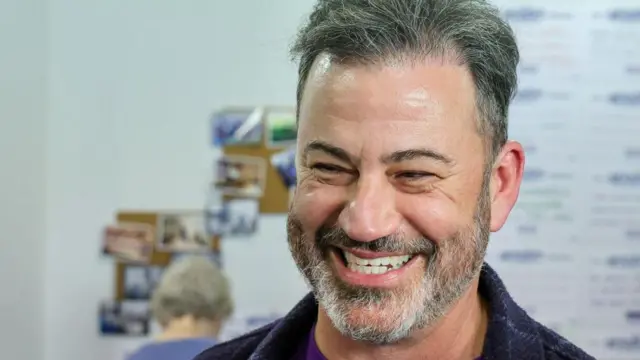 Jimmy Kimmel, sonriente, ante un fondo difuminado en el que se ve la parte trasera de la cabeza de una persona