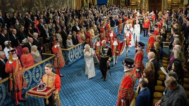 Queen Elizabeth II is accompanied by her son Britain"s Prince Charles, Prince of Wales walk behind the Imperial State Crown as they proccess through the Royal Gallery during the State Opening of Parliament in the Houses of Parliament in London on October 14, 2019.