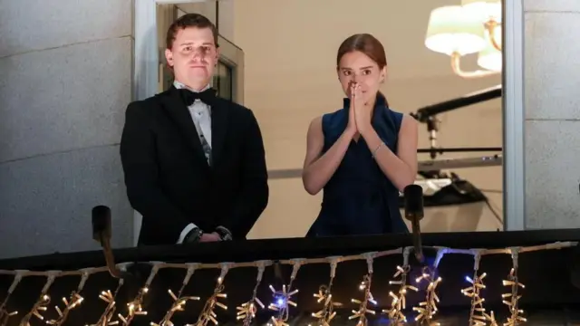 黒のタキシードを着たマチャド氏の息子と、黒のノースリーブ・ワンピースを着たマチャド氏の娘が、ホテルのバルコニーに並んで立ち、通りに集まった人々を見ている