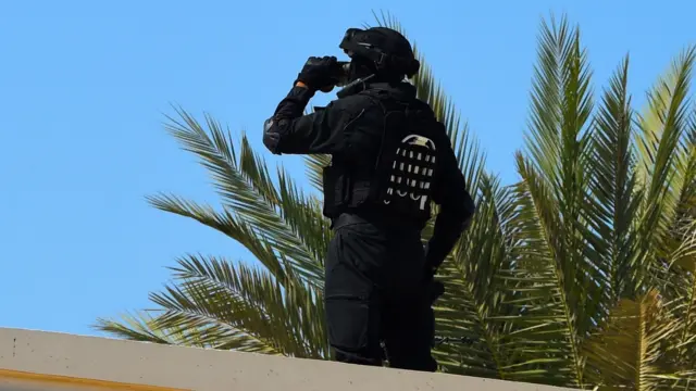 Un guardia de seguridad bahreiní se encuentra sobre una azotea junto a una palmera en un día soleado. Se lleva la mano al rostro y viste un uniforme negro con casco negro.