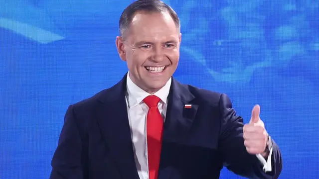 Prezydent Polski Karol Nawrocki - mężczyzna w średnim wieku, o krótko ostrzyżonych włosach - szeroko się uśmiecha i pokazuje kciuk w górę. Jest ubrany w granatową marynarkę z przypinką w kształcie polskiej flagi, białą koszulę i czerwony krawat. W tle jasnoniebieski ekran lub baner. 