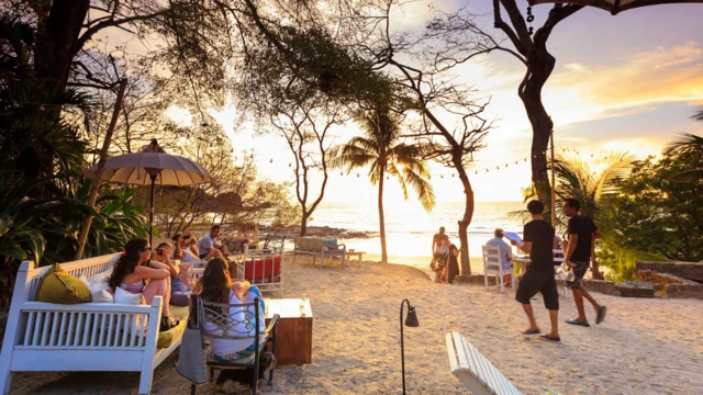Na małej plaży rozstawiono krzesła i ławki, na których siedzi kilkanaście osób, głównie młodych. Na drugim planie widać kilka drzew, w tym palmowych. Dalej morze i niebo o zachodzie słońca. 