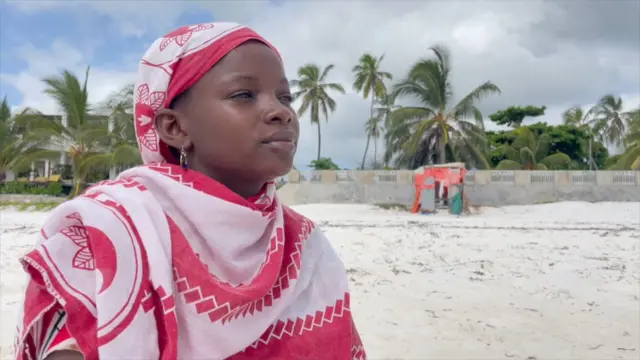 Uma mulher jovem, negra, com um lenço corem quem apostar hoje futebolrosa e vermelho sobre o peito e cabeçaem quem apostar hoje futeboluma praiaem quem apostar hoje futebolareia branca. Ao fundo, coqueiros