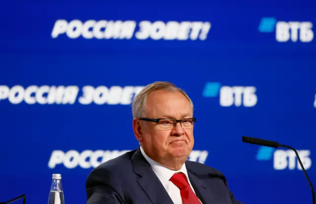 俄罗斯国有银行VTB的董事长科斯京戴着眼镜,打着红色领带,坐在一个带有俄文文字和VTB标志的亮蓝色背景前