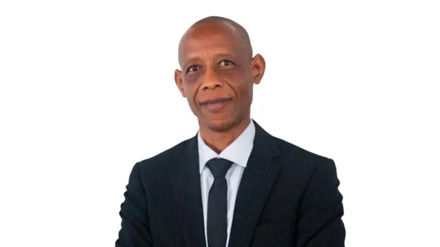 Ifoto ngufi igaragaza Profeseri Emile Bienvenu yambaye ikositimu y'umukara, ishati y'umweru na karuvati y'umukara. Arareba muri 'camera'.