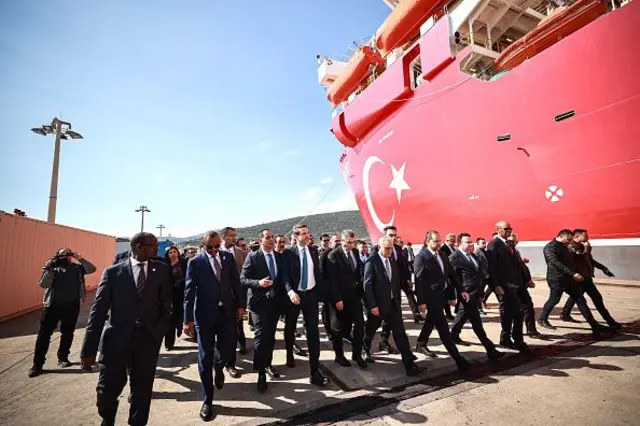 Resmi giyimli onlarca erkek, Türk bayrağı deseniyle kaplı gemi gövdesinin yanında yürüyor.