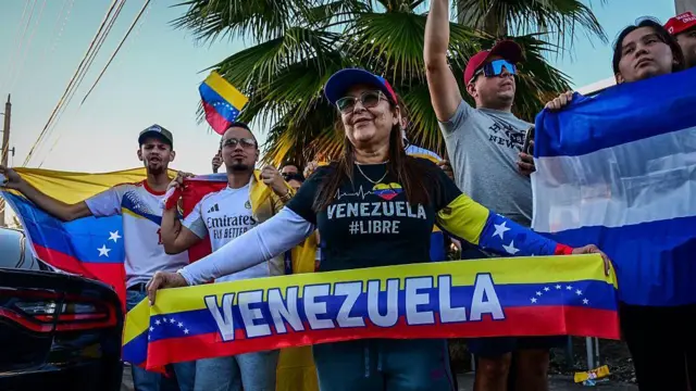 Un pequeño grupo de personas celebrando con banderas y pancartas venezolanas en Doral, un suburbio de Miami. Están afuera y hay palmeras al fondo.