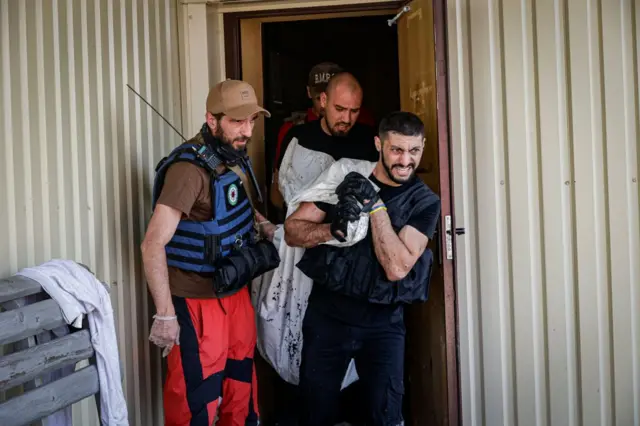 Кілька чоловіків у захисному спорядженні виходять із будівлі, несучи загорнуте в біле покривало тіло.