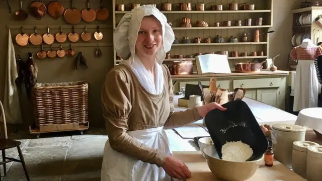 Una joven con un traje de la época de la regencia en una cocina. Lleva una blusa beige de manga larga con una blusa blanca debajo, un delantal blanco y un gran sombrero blanco. Sostiene una bandeja negra y vierte harina en un tazón beige.