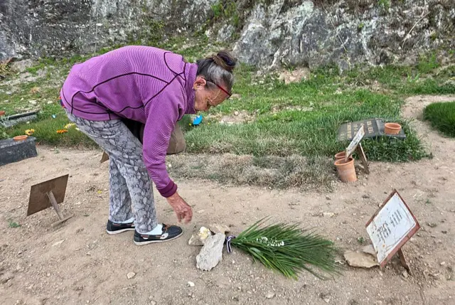 Carmen Nava deposita flores en la tumba de su hijo. Es un suelo de tierra donde la única identificación es una placa metálica con lo que parece un papel encima donde pone el nombre de su hijo. Ella está agachada, tras depositar un ramo que ha dejado pillado con varias piedras. Es una mujer visiblemente mayor, encorvada.