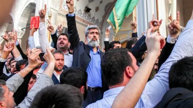 Alparslan Kuytul, etrafında çok sayıda kişiyle birlikte bir caminin önünde işaret parmaklarını yukarı kaldırmış