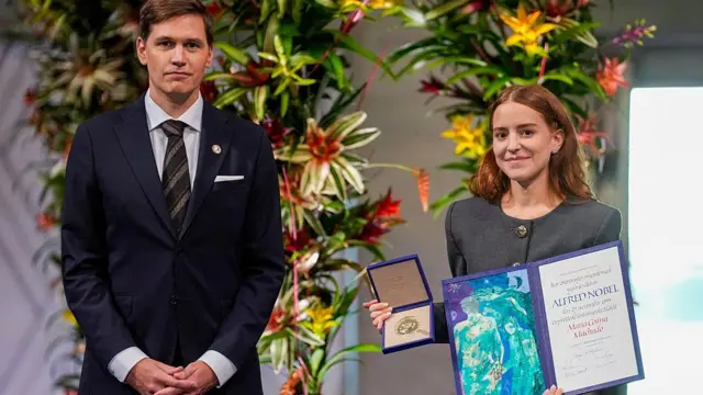 Ana Corina Sosa Machado, quien viste un traje gris, sostiene el premio nobel de la paz y un certificado de la organización del nobel. Está de pie junto a Jorgen Watne Frydnes, presidente del Comité Noruego del Nobel, quien viste de traje y corbata.