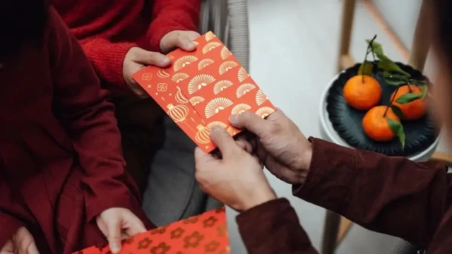 Два красных конверта, украшенные один золотистым орнаментом в виде китайских фонариков, другой орнаментом в виде веером. Их с двух сторон держат два человека. В кадре только руки, но видно, что один из них постарше, другой помладше. Справа на тарелке лежат три мандарина.