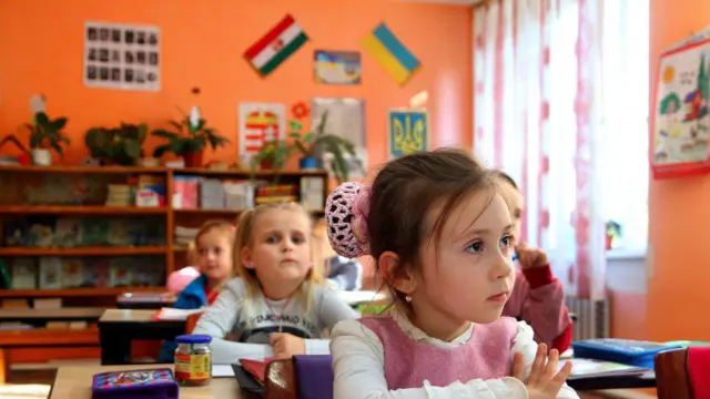 Шкільний клас, прикрашений плакатами та прапорцями України та Угорщини. Маленька дівчинка сидить за партою, склавши руки. Позаду неї - інші діти. 