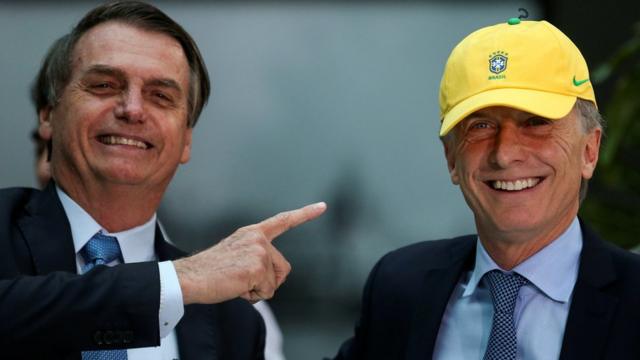 Bolsonaro aponta para Macri, que veste boné da seleção brasileira; ambos sorriem