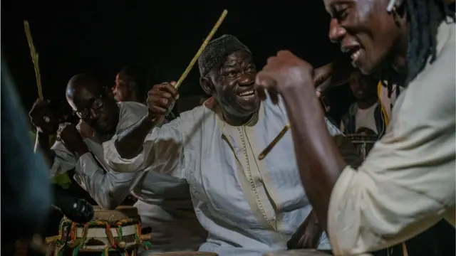 Babou Ngom playing a drum-like instrument with a drum stick in the air. He is smiling. Others around him are performing and visibly having a good time.