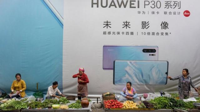 Women sell vegetables in front of a billboard advertising smartphones for China's Huawei Technologies Co., on June 1, 2019 in Mangshi, Yunnan Province, southwestern China