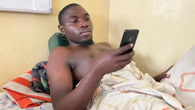 Mundeke Kandundao est allongé sur un lit d'hôpital, torse nu, en train de regarder son téléphone portable.