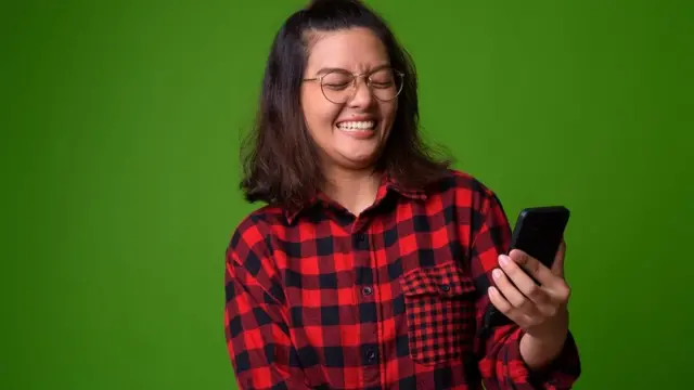 Jeune femme avec une chemise à carreaux rouge et noir. Elle rit et tient un smarphone. Fond vert vif.