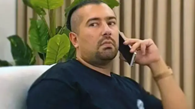 Rawa Majid siedzi na krześle i rozmawia przez telefon. Ma zaczesane do tyłu, wygolone po bokach włosy i kozią bródkę. Patrzy w bok ze skupionym wyrazem twarzy. 