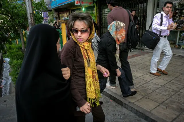 ضابطة من شرطة الأخلاق الإيرانية تمسك بذراع امرأة ترتدي نظارة شمسية في أحد شوارع طهران، كان شعرها غير مُغطى.