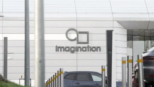 现代白色建筑外观，墙上印有 Imagination Technologies 标志，图案由圆形与方形组成，位于“Imagination”字样上方。前景排列多个防撞柱，入口附近可见停放的车辆。