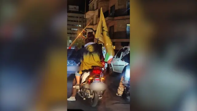 تصویر دو زن سوار بر موتور در تجمع‌های حکومتی شبانه، در حالی که پرچم حشد شعبی و جمهوری اسلامی در دست دارند.