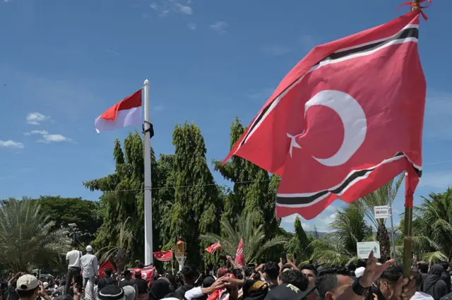 Seorang pria (kiri atas) memanjat tiang bendera saat ia berupaya mengibarkan bendera Gerakan Aceh Merdeka (GAM) bersandingan dengan bendera nasional Indonesia dalam sebuah unjuk rasa di luar Masjid Raya Baiturrahman, Banda Aceh, Provinsi Aceh, pada 15 Agustus 2026.