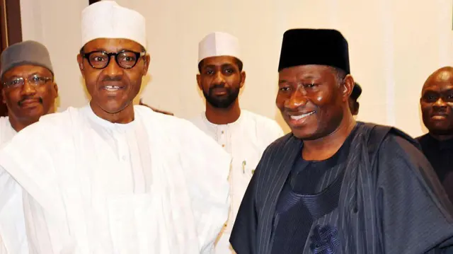 Muhammadu Buhari, in white traditional Nigerian clothes, and Goodluck Jonathan, in dark blue traditional clothes, stand next to each other smiling.