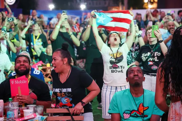 Fanáticos viendo el show de medio tiempo de Bad Bunny en el Super Bowl. Una mujer sostiene la bandera de Puerto Rico