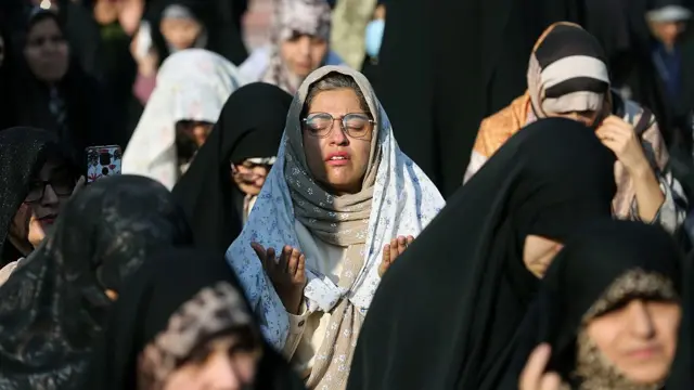 Tahran’da dua eden örtülü kadınlar görülüyor. Çoğunda siyah başörtüsü var. Ortadaysa gözlük takan bir kadın gözleri kapalı şekilde dua ederken ellerini kaldırmış olarak görülüyor.
