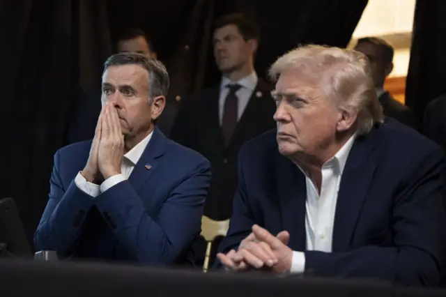 Ratcliffe con las dos manos juntas frente a su rostro, y Trump con cara seria y las manos sobre la mesa, una encima de la otra.