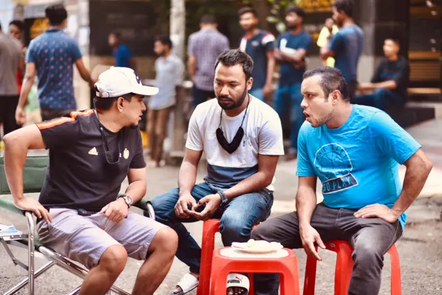 ব্যাচেলর পয়েন্ট নাটকের একটি দৃশ্য যেখানে তিনজন ব্যক্তি বসে কথা বলছেন, তাদের সামনে একটি মোড়ার ওপর প্লেটে কিছু পিঠা। পেছনে দাঁড়িয়ে আছেন কয়েকজন