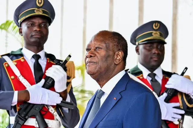 Le président Alassane Ouattara en costume bleu, chemise blanche et cravate bleue, regarde ailleurs. Deux hommes en uniforme militaires portant des armes sont en arrière-plan