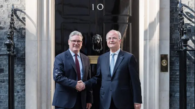 کی‌یر استارمر و محمد مصطفی روی پله‌های خیابان داونینگ شماره ۱۰ ایستاده‌اند و دست‌هایشان را محکم به هم می‌فشردند. در مشکی و براق اقامتگاه با شماره ۱۰ درست پشت سر آنهاست. هر دو مرد کت و شلوار سورمه‌ای پوشیده‌اند. استارمر کراوات قرمز تیره و مصطفی کراوات آبی روشن دارد. نور خورشید روی در و چارچوب‌های سفید در می‌تابد