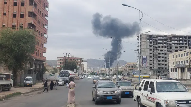 حركة السير ومواطنون في شارع رئيسي وخلفهم تصاعد دخان عقب غارة إسرائيلية في صنعاء.