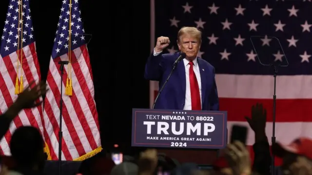 Donald Trumpo, um homem branco idosoxbet99 fazer cadastroterno e cabelo loiro, durante um evento com apoiadores; ao fundo, bandeira dos EUA