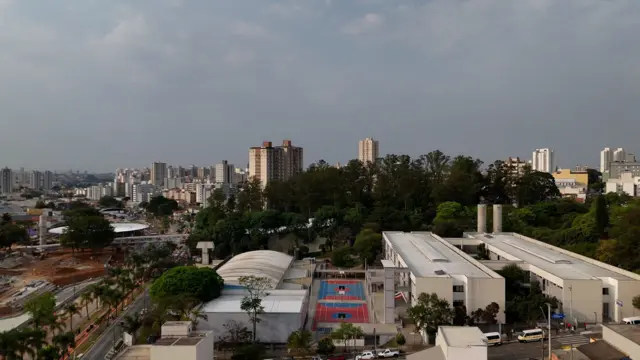 Vista aérea de quadra esportiva ladeada por edifícios, com prédios e árvores ao fundo