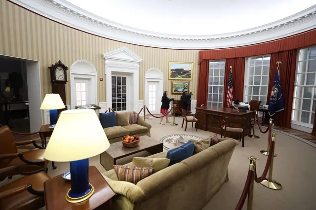 Sillones tapizados, lámparas y muebles de madera oscura llenan una réplica del Despacho Oval, con partes del espacio delimitadas por cuerdas de terciopelo rojo. Dos personas muestran el espacio.