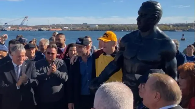 Chuck Wepner mengenakan topi kuning, berpose dengan patung yang didirikan untuk menghormatinya di New Jersey.