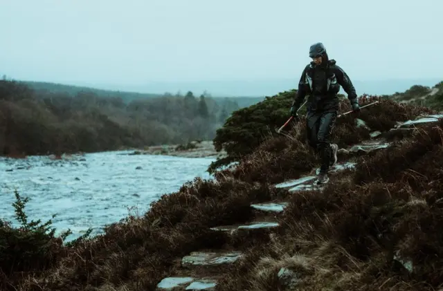 Una corredora solitaria vestida de verde oscuro baja unas escaleras junto a un gran río con un bosque a lo lejos.