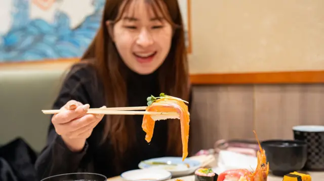 A close-up of a happy young Asian woman in a Japanese restaurant, lifting a freshly presented salmon nigiri with chopsticks from a plate full of a variety of sushi.