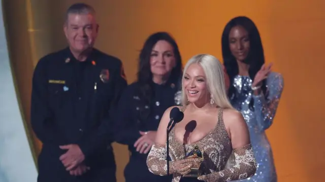 Бейонсе у микрофона со статуэткой «Грэмми», за ней стоят двое пожарных и ведущая церемонии.