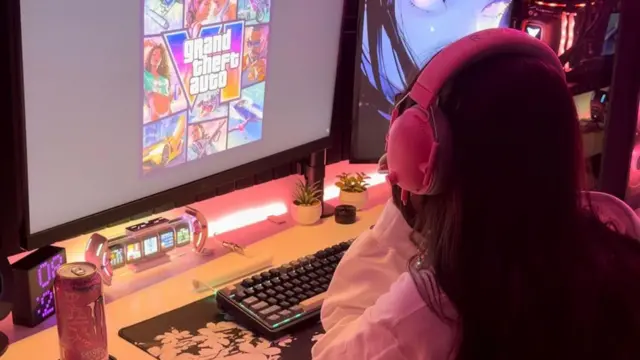 一個從肩後視角拍攝的畫面，顯示一名女子——內容創作者Alogirlx——正注視著她的電腦螢幕，螢幕上顯示《GTA 6》的藝術圖像與標誌。她戴著粉紅色耳機，而她擺放鍵盤的桌面則被明亮的粉紅色LED燈照亮。