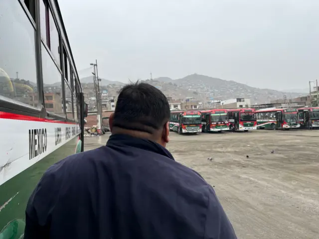 Toño, un hombre con una chaqueta azul, está parado al lado de un bus en un gran lote. Hay más buses al fondo del lote y detrás, se ve una montaña con barrios de invasión.