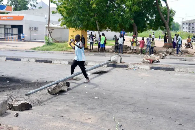 Un jeune enjambe un barrage de fortune sur une route. De l'autre c^té de la chaussée un groupe de gens se tient debout