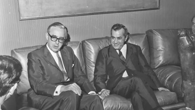 Uma fotografia em preto e branco mostra dois homens de terno sentados em um sofá, conversando, enquanto outras pessoas aparecem parcialmente ao redor. A cena parece registrar um encontro formal em um ambiente interno.